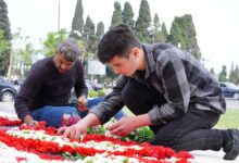 Two men placing red and white flowers at an outdoor memorial on a sunny day.
