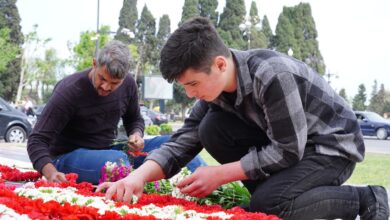 Two men placing red and white flowers at an outdoor memorial on a sunny day.