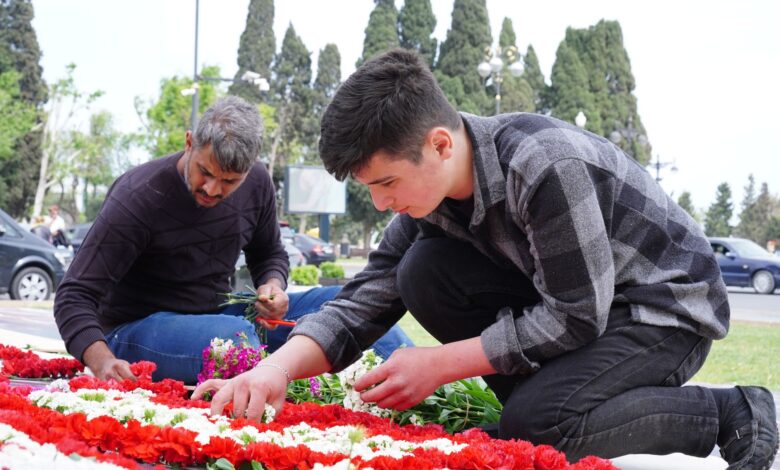 Two men placing red and white flowers at an outdoor memorial on a sunny day.