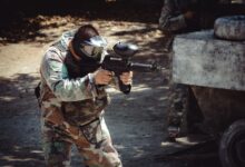 Man in camouflage gear aiming a paintball gun during an outdoor game.