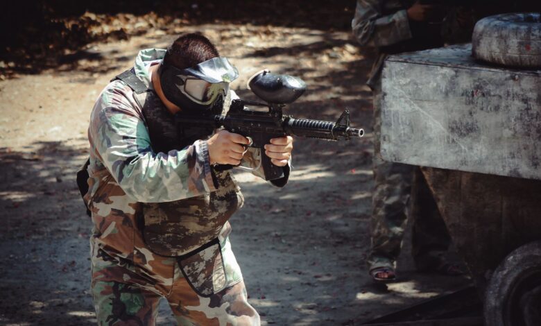 Man in camouflage gear aiming a paintball gun during an outdoor game.