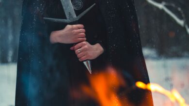 A cloaked figure holds a sword amidst snow and flames, evoking mystery.