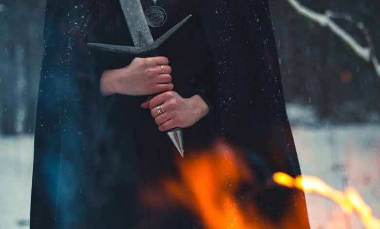 A cloaked figure holds a sword amidst snow and flames, evoking mystery.