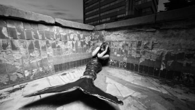 Black and white photo of a merperson posing in an urban corner, evoking mystery and fantasy.