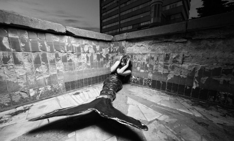 Black and white photo of a merperson posing in an urban corner, evoking mystery and fantasy.