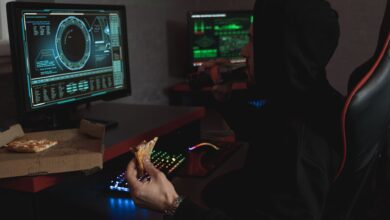 Hacker in a black hoodie eating pizza and drinking soda while analyzing data on multiple computer screens.