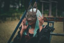 A woman with green hair wearing an ornate rabbit mask poses outdoors, exuding a mysterious vibe.