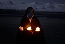 Unrecognizable person in plague mask standing in front of snowy hills at night and holding burning candles while looking at camera