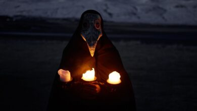 Unrecognizable person in plague mask standing in front of snowy hills at night and holding burning candles while looking at camera