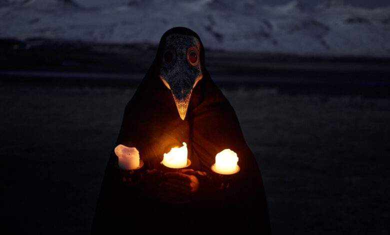 Unrecognizable person in plague mask standing in front of snowy hills at night and holding burning candles while looking at camera