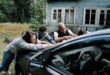 A group of zombies surround a car in front of an old, abandoned house.