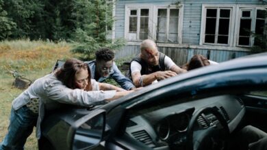 A group of zombies surround a car in front of an old, abandoned house.