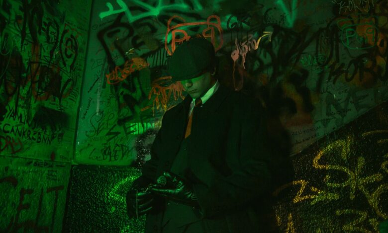 Stylish man in a trench coat and hat standing against vibrant graffiti wall, exuding retro elegance.
