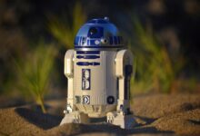 Close-up of an R2-D2 toy standing on sand, set against a green background in bright daylight.
