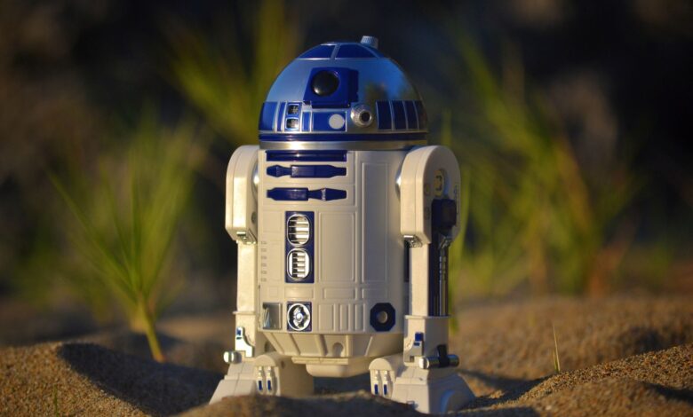 Close-up of an R2-D2 toy standing on sand, set against a green background in bright daylight.