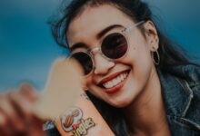 Smiling woman in sunglasses and denim jacket holding Pringles chip outdoors.