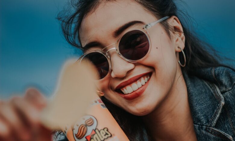 Smiling woman in sunglasses and denim jacket holding Pringles chip outdoors.