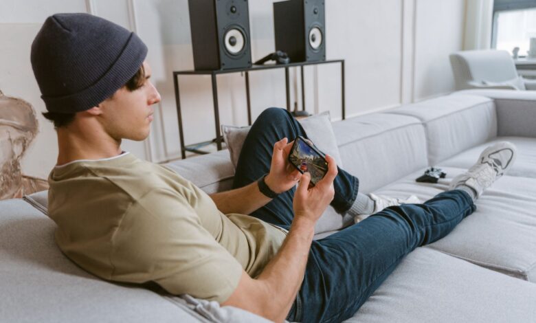 Casual young man playing mobile games on sofa with speakers in the background.