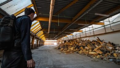 An urban explorer inspects the interior of an abandoned warehouse filled with debris.