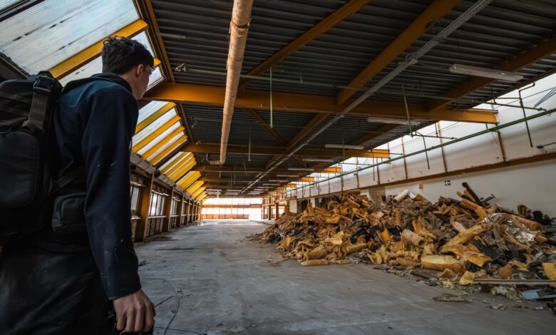 An urban explorer inspects the interior of an abandoned warehouse filled with debris.