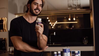 Smiling man holding a supplement bottle in an indoor kitchen setting with warm lighting.
