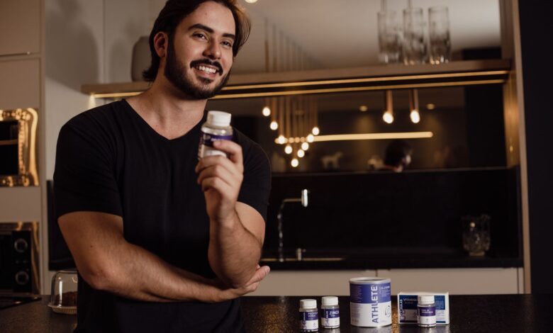 Smiling man holding a supplement bottle in an indoor kitchen setting with warm lighting.