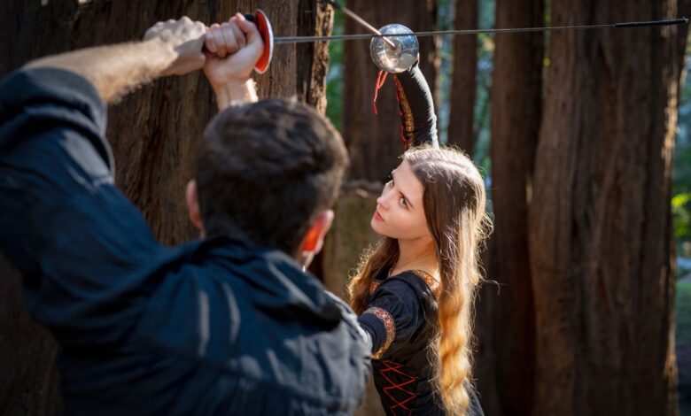 A man and woman engage in a historical sword duel in the woods.