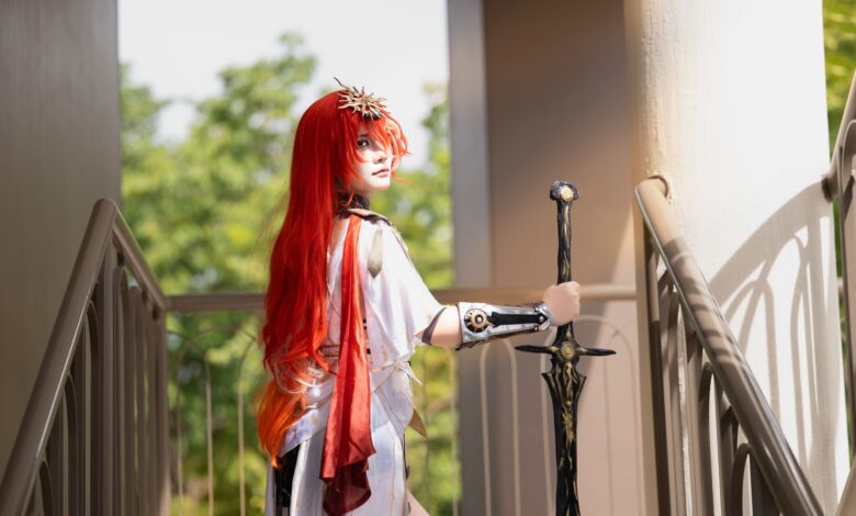 Cosplayer with striking red hair holds a sword on a sunlit stairway, dressed in elaborate costume.