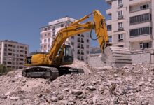 Yellow excavator working on urban construction site amidst rubble and buildings in Kahramanmaraş, Türkiye.