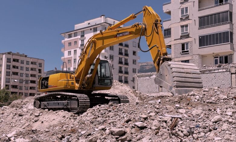 Yellow excavator working on urban construction site amidst rubble and buildings in Kahramanmaraş, Türkiye.