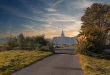 A serene road surrounded by greenery leading to a distant church under a colorful sunset sky.