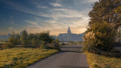 A serene road surrounded by greenery leading to a distant church under a colorful sunset sky.