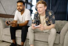 Two friends sitting on a sofa, playing video games together at home.