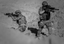 Two soldiers in camouflage and gear in a desert setting, engaged in a tactical action.