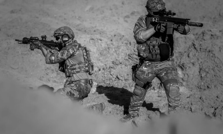 Two soldiers in camouflage and gear in a desert setting, engaged in a tactical action.