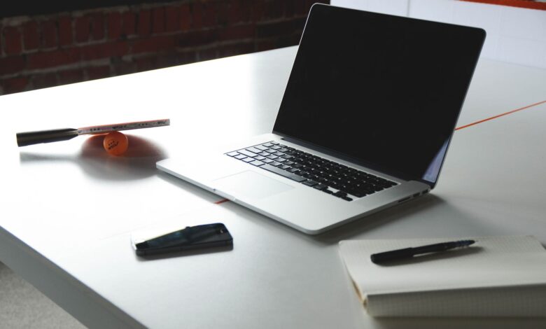 A sleek workspace featuring a laptop, notebook, smartphone, and table tennis paddle on a desk.
