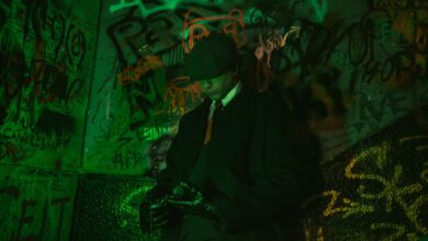 Stylish man in a trench coat and hat standing against vibrant graffiti wall, exuding retro elegance.