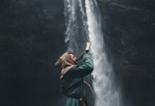 A warrior in a hooded cloak standing by a waterfall in Iceland, holding a horn.