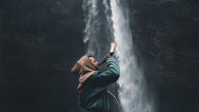 A warrior in a hooded cloak standing by a waterfall in Iceland, holding a horn.