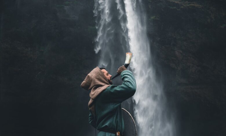 A warrior in a hooded cloak standing by a waterfall in Iceland, holding a horn.