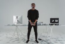 A handcuffed man stands in a room with scattered money, a computer, and a briefcase.