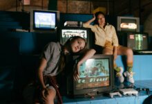 Two women in casual outfits immersed in retro gaming with vintage televisions and game consoles.