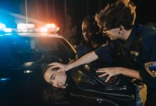 Two police officers arrest a suspect during a nighttime operation with flashing lights.