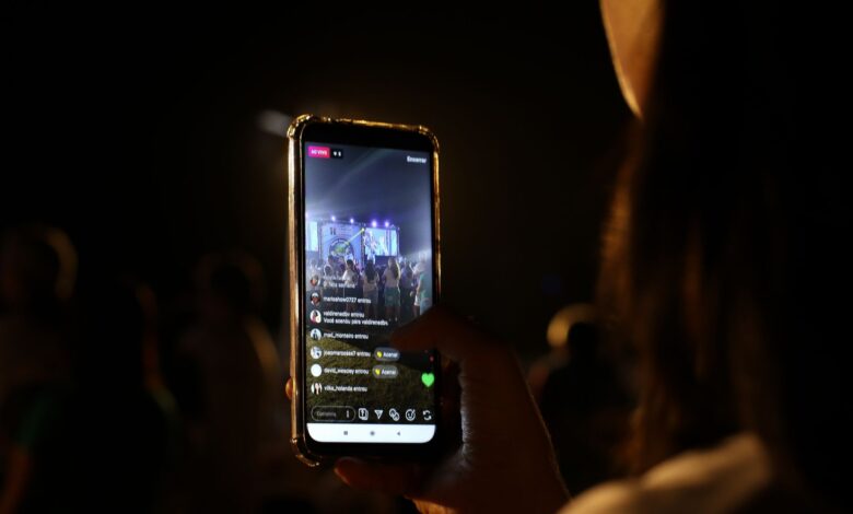 A woman live streams a concert using her smartphone at night. Vibrant stage in view.