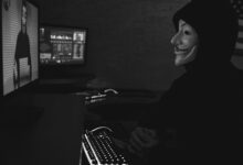 Monochrome image of a hacker in a Guy Fawkes mask working on a computer in a dimly lit room.