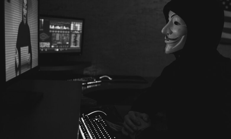 Monochrome image of a hacker in a Guy Fawkes mask working on a computer in a dimly lit room.