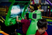 Unrecognizable child having fun with bright car racing simulator while turning steering wheel near serious ethnic woman and funny brother in shopping center near playground