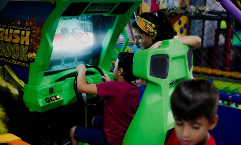Unrecognizable child having fun with bright car racing simulator while turning steering wheel near serious ethnic woman and funny brother in shopping center near playground