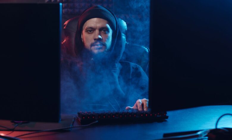 Dramatic scene of an adult man in a hoodie using a computer, surrounded by smoke in a dimly lit room.