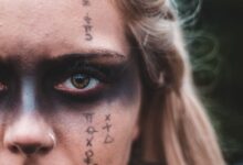 Closeup crop blond with black painted mask on eyes and symbols on skin looking at camera